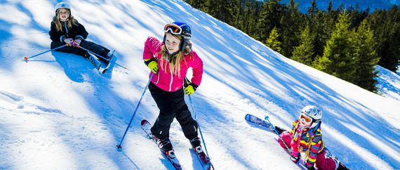 Mädchen in bunter Skikleidung und mit Helmen auf Abfahrtsskiern, die an einem stark verschneiten Berghang im Schnee liegen bzw. auf Skiern stehen und nach oben schauen