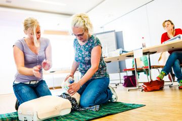 Das Bild zeigt einen Workshop, in dem zwei Frauen am Boden mit einer Puppe knien.