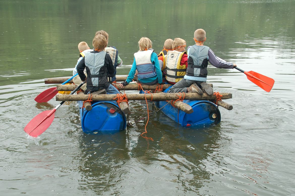 Eine Gruppe von Schülern auf einem selbstgebauten Floß auf einem See. Die Kindergruppe wird von hinten gezeigt.