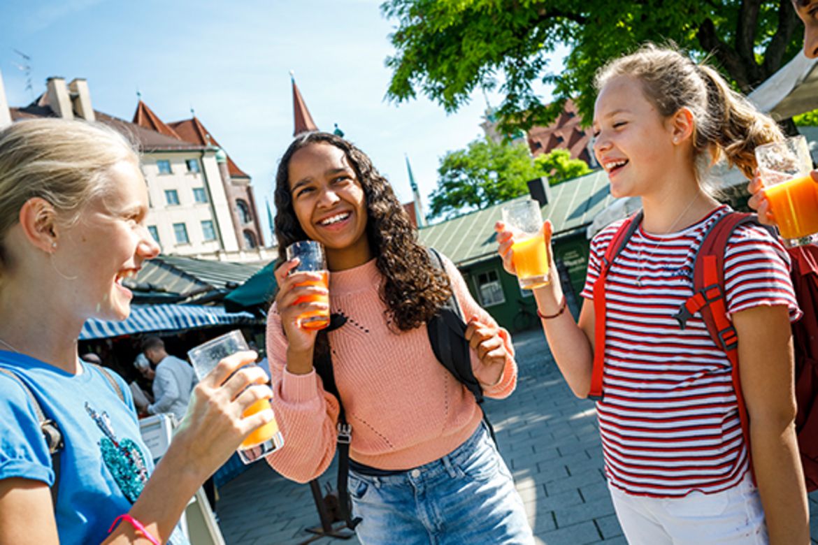 Drei Kinder auf dem Viktualienmarkt in München, jeweils mit einem Glas Fruchtsaft in der Hand.