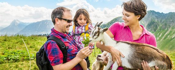 Eine Familie in der freien Natur auf einer Bergwiese. Die Frau hat eine Ziege auf dem Arm, der Mann hält ihr Blumen zum Fressen hin.