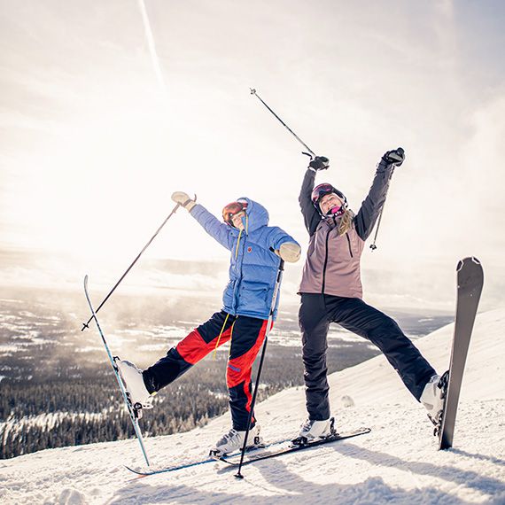 Zwei jugendliche Personen stehen freudig auf einem Berg in Skikleidung. Ihre Skistöcke halten sie in die Luft und stellen einen Ski auf dem Schnee ab.