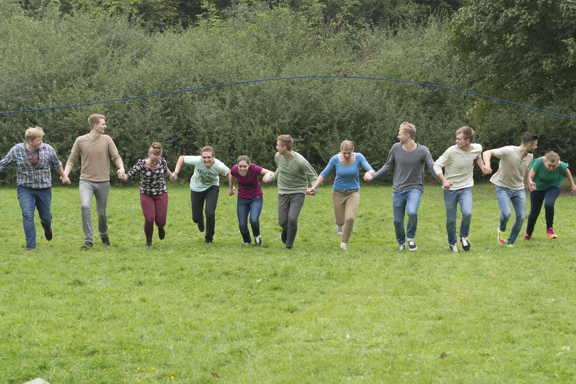 Elf junge Menschen laufen, sich an der Hand haltend, lachend und geduckt unter einem langen Springseil hindurch