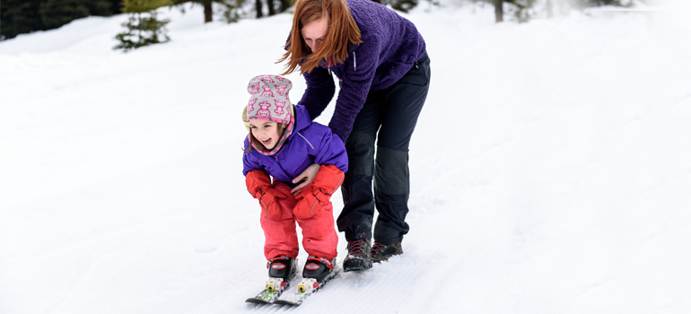 Ein junges Mädchen in farbenfroher Skiausrüstung und auf Skiern wird von ihrer Mutter angeschubst und lacht. Im Umfeld erkennt man eine verschneite Landschaft.