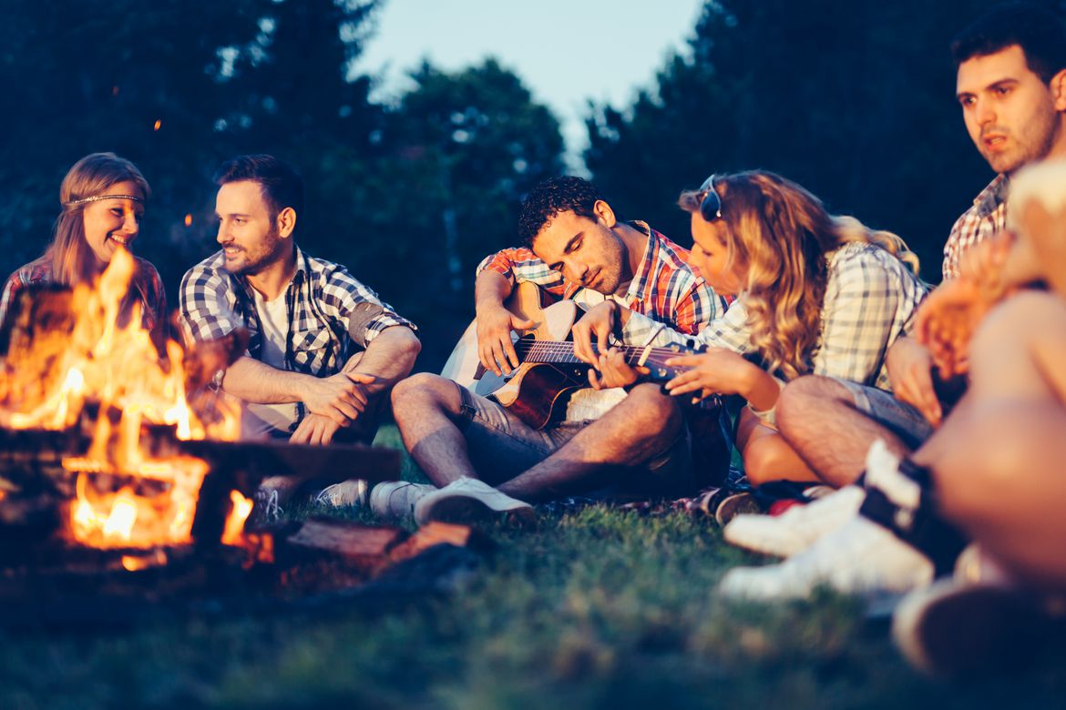 Eine Gruppe junger Erwachsener sitzt gemeinsam am Lagerfeuer. Einer spielt eine Gitarre.