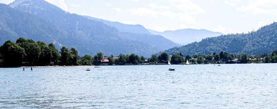 Blick über einen bayerischen See, im Hintergrund bewaldete Hügel, auf dem See einige Wassersportler.