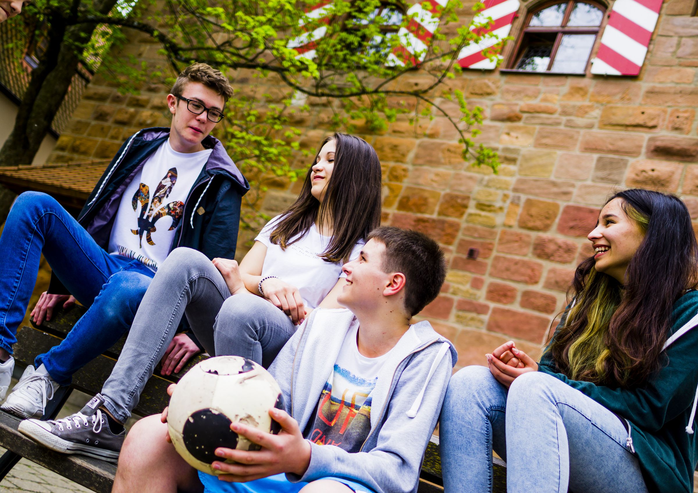 Vier Jugendliche chillen auf einer Bank, einer von ihnen hat einen Fußball in der Hand.