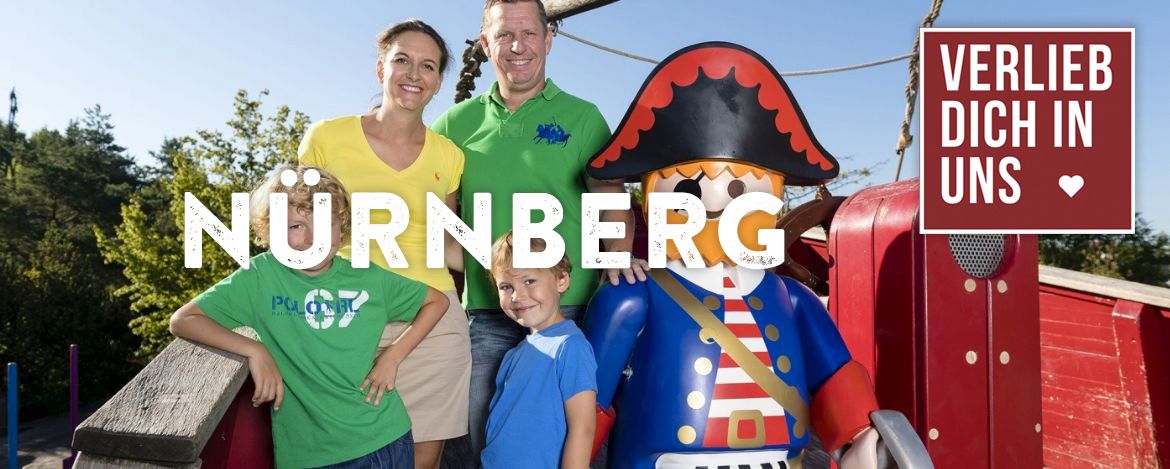 Eine Gruppenaufnahme einer jungen glücklichen Familie neben einer lebensgroßen Piraten-Playmobil Figur. Das alles befindet sich im Playmobil Funpark in der Nähe von Nürnberg. Die Beschriftung ist Nürnberg und der Hinweis auf das Verlieb dich in uns Angebot befindet sich in der rechten Ecke.