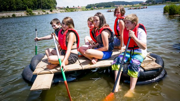 sechs Kinder sitzen auf einem selbstgebauten Floß und paddeln