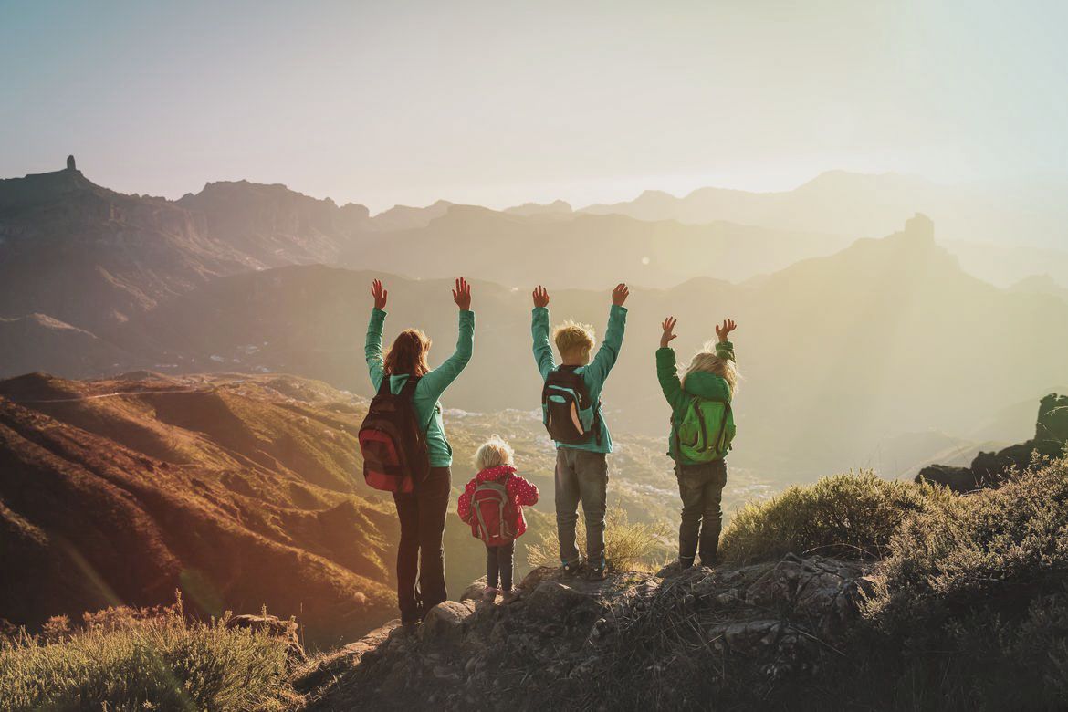 Eine Frau steht mit drei Kindern auf einem Berg. Alle haben die Hände nach oben hochgestreckt. Das Foto selbst zeigt eine Berglandschaft in Dämmerungsstimmung.