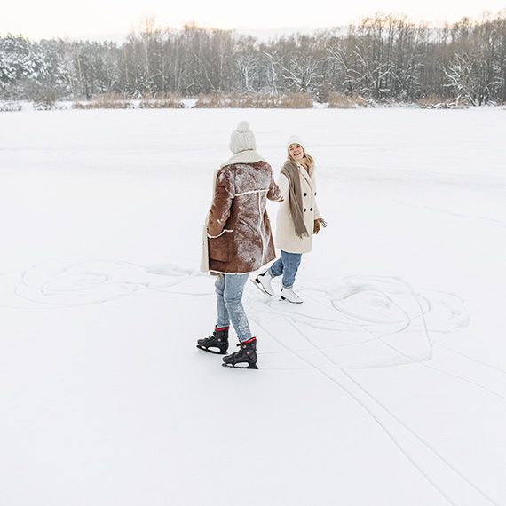 Zwei Frauen beim Schlittschuhlaufen auf einem zugeschneiten See, die sich dabei an den Händen halten