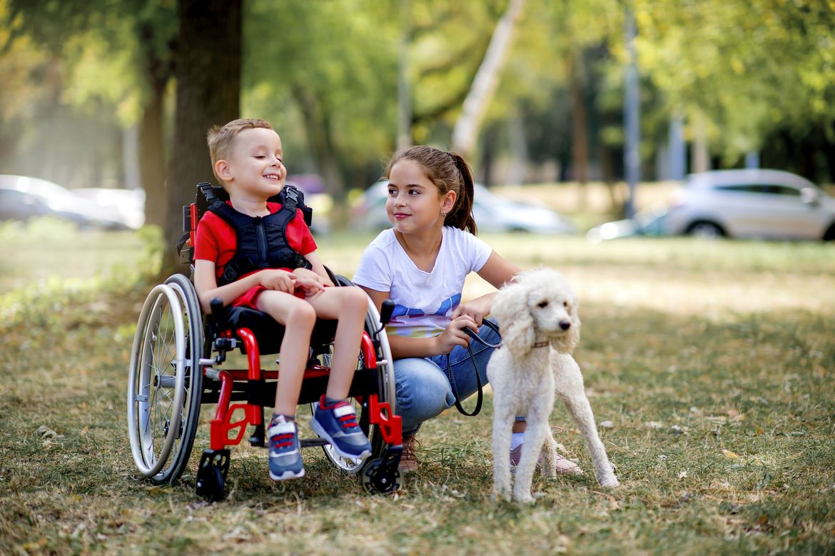Ein Junge im Rollstuhl, ein Mädchen kniet mit einem Hund daneben