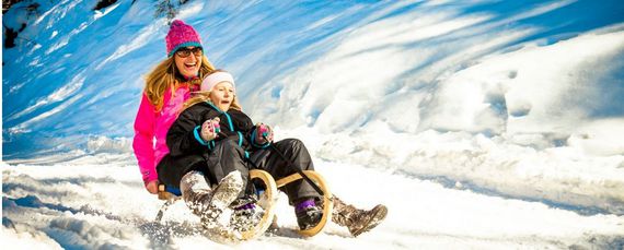 Eine Mutter und ihr Kind rodeln quietschvergnügt einen Rodelhang herunter. Beide tragen warme Skisachen.