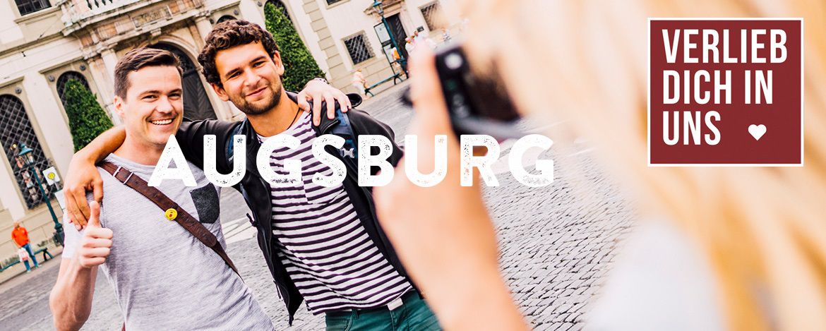 Zwei junge Männer lassen sich von einer Frau fotografieren. Im Hintergrund ist eine historische Hausfassade zu erkennen. Das Bild wird überlagert von den Schriftzügen "Verlieb dich in uns" und "Augsburg".