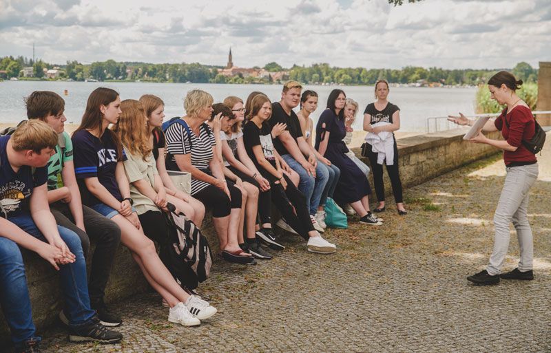 Die Klasse sitzt auf einer niedrigen Mauer am Schwedtsee. Im Hintergrund ist der Ort Fürstenberg an der Havel zu erkennen.