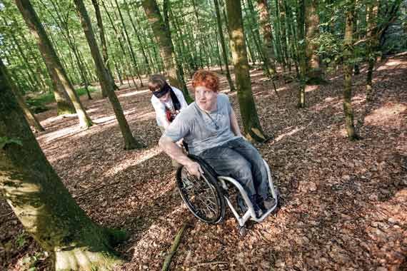 Ein Schüler mit verbundenen Augen hilft einem Schüler im Rollstuhl durch den Wald zu kommen. 