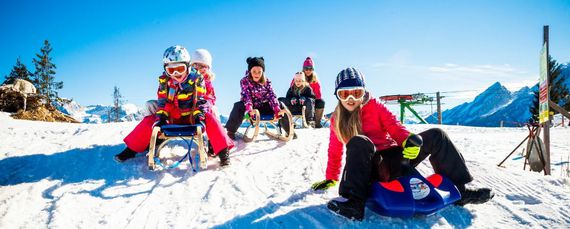 Kinder in bunten Skisachen rodeln einen Berghang hinunter.