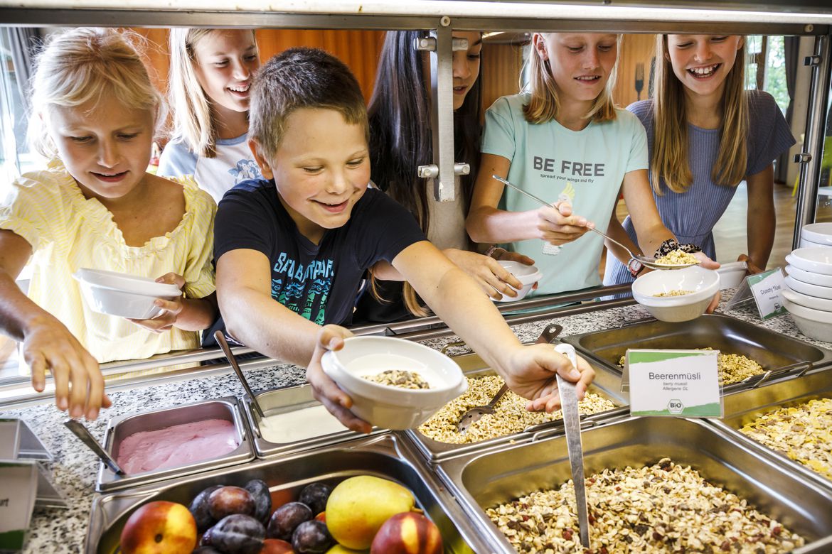Sechs Schulkinder stehen am Buffet, bei dem eine Vielzahl von Müsli, Joghurt und Obst zu sehen ist. Die Kinder bedienen sich mit hungrigen Augen
