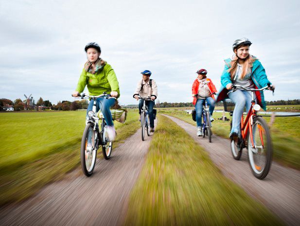 Auf dem Bild fahren im Vordergrund zwei jüngere Frauen mit einem Helm. Im Hintergrund sieht man zwei altere Personen auf dem Fahrrad.