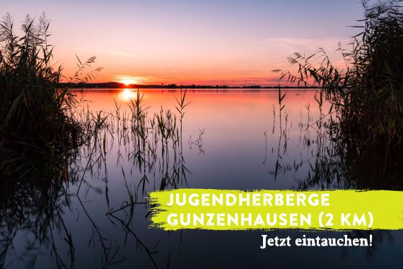 Blick über den Altmühlsee im Sonnenuntergang. Das Foto wird überlagert von dem Schriftzug "Jugendherberge Gunzenhausen - jetzt eintauchen!"