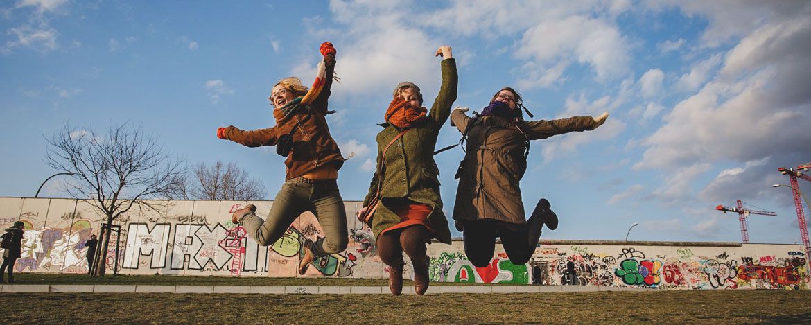 Drei junge Frauen springen in die Luft. Im Hintergrund sieht mal ein Teilstück der Berliner Mauer