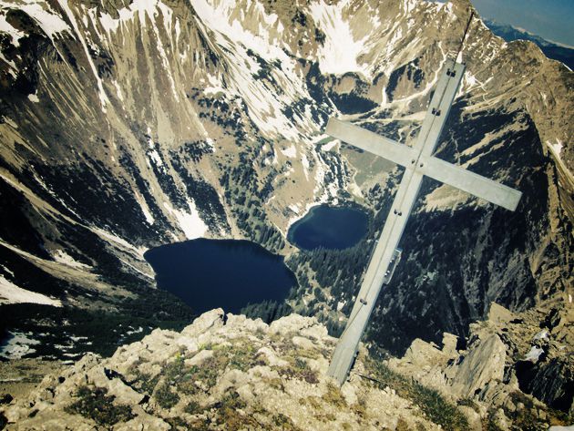 Blick hinunter zu einem Bergsee, im Vordergrund ein einfaches Kreuz, an dem eine metallene Antenne befestigt ist