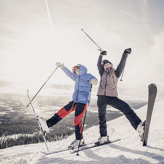 Zwei jugendliche Personen stehen freudig auf einem Berg in Skikleidung. Ihre Skistöcke halten sie in die Luft und stellen einen Ski auf dem Schnee ab.