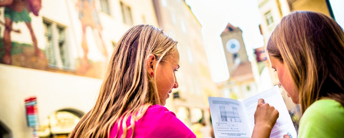 Zwei Kinder informieren sich io einem gedruckten Faltblatt über ein im Hintergrund zu erkennendes Gebäude