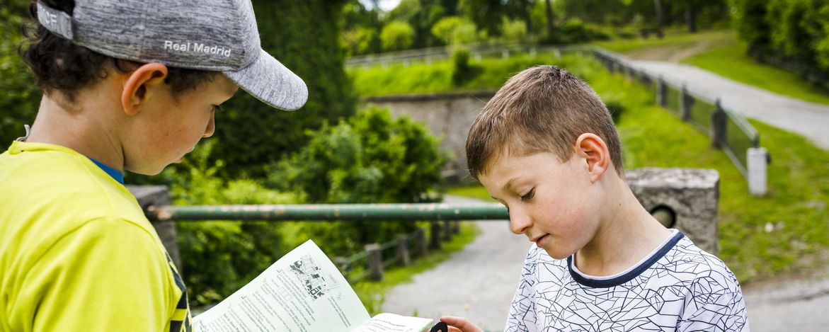 Zwei Kinder studieren im Freien eine gedruckte Beschreibung. Ein Kind hält einen Kompass bereit.