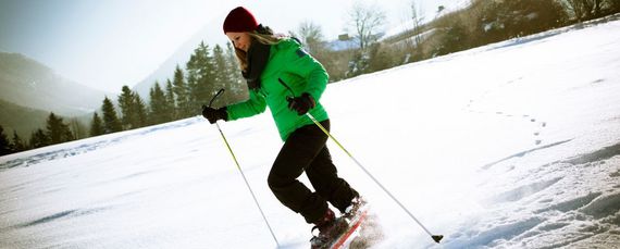 Eine junge Frau in grüner Skijacke auf Schneeschuhen in verschneiter Winterlandschaft unterwegs